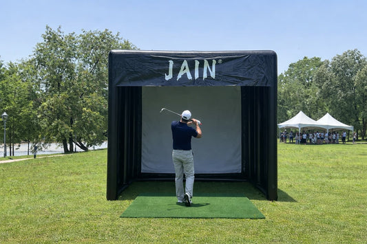 A professional JAIN inflatable golf simulator tent set up at an outdoor charity tournament with a golfer swinging and event tents in the background.