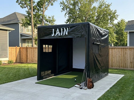A quiet, professional home golf simulator setup on a backyard patio, showing an inflatable JAIN tent with a green hitting mat ready for use.