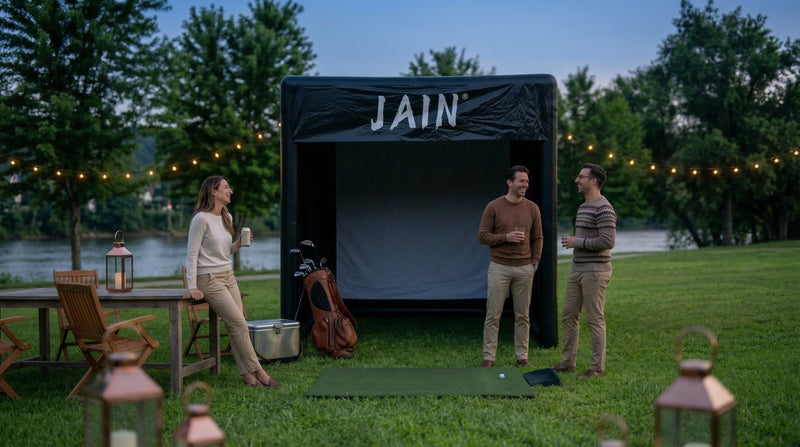 Three friends socialize at dusk in front of a "JAIN" branded inflatable outdoor golf simulator by a river.