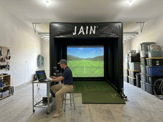 A golfer relaxing at a laptop stand next to a funjain inflatable golf simulator enclosure inside a spacious residential garage, featuring synced course projections on both the laptop and the large impact screen.