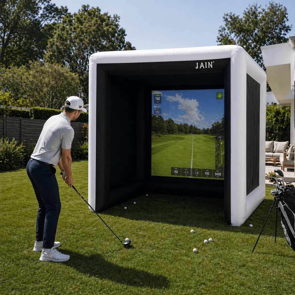 Golfer practicing with a JAIN inflatable golf simulator tent on a backyard lawn with simulator screen and golf balls
